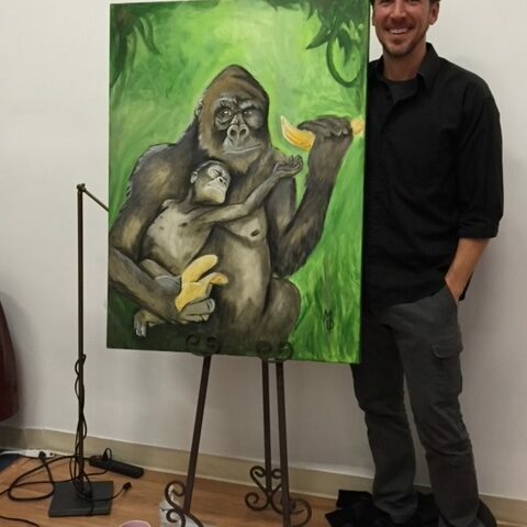 This is an image of an artist next to his completed work, the painting is of a gorilla and its infant. The painting has a green background, with a gorilla holding a banana while a baby gorilla reaches out towards it.